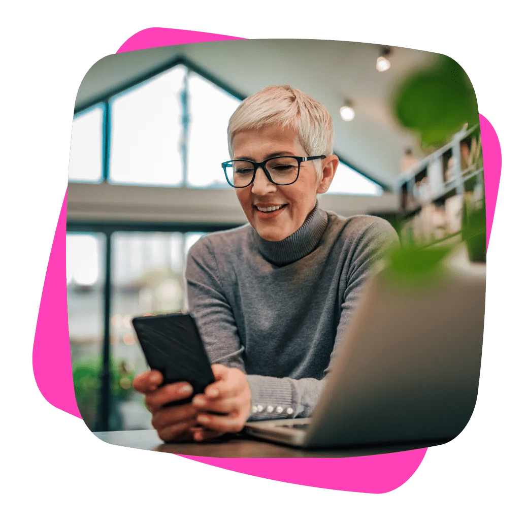 Older lady with eyeglasses, using her phone with laptop on the side
